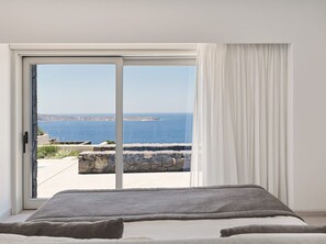 A bedroom with a large sliding glass door leading to a balcony with a view of the sea and a rocky shoreline. - Villa Elythiona, Divine Sea Views, Private Pool, Furnished Terrace, Sanrotini (Oia, Santorini)