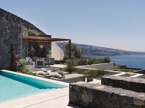 A poolside area with a stone wall, outdoor seating, and a view of the sea. - Villa Elythiona, Divine Sea Views, Private Pool, Furnished Terrace, Sanrotini (Oia, Santorini)