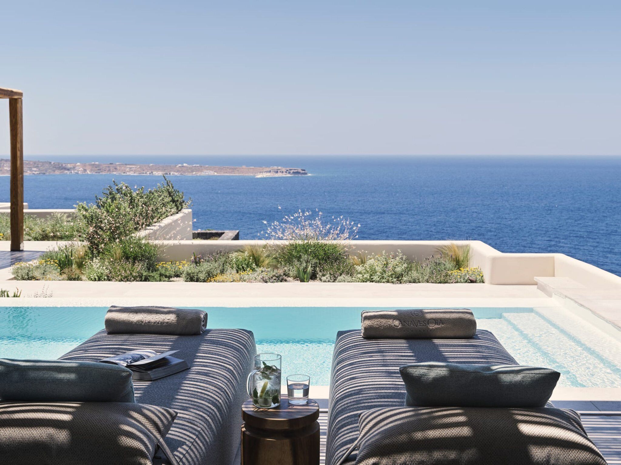 A poolside area with two lounge chairs, a small table with a glass and a bottle, and a view of the sea.