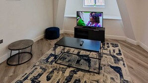 Un salon avec un téléviseur posé sur un meuble, un petit pouf rond, une table basse et un tapis à motifs.