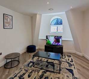 Een woonkamer met een televisie op een dressoir, een klein rond voetenbankje, een salontafel en een kamerbreed tapijt.