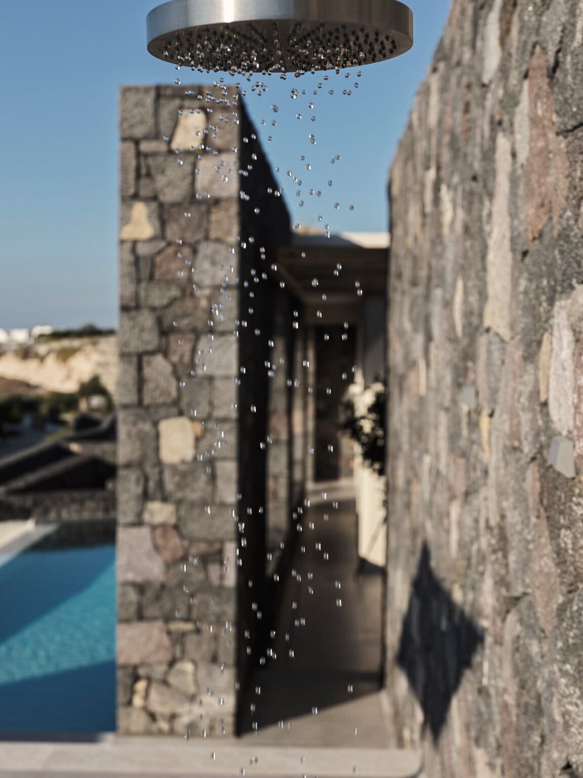 Eine moderne Außendusche mit Wassertropfen, einer Steinsäule und einem Pool im Hintergrund.