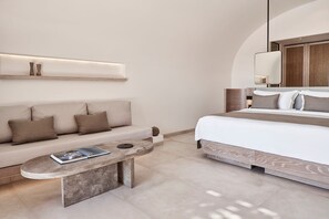 A modern hotel room with a sofa, a small round table with a book, a bed with a wooden headboard, and a wall-mounted shelf with decorative items. - Avenithion Suite, Divine Sea Views, Private Pool, Furnished Terrace, Sanrotini (Santorini)