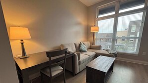 A modern living room with a sofa, a coffee table, a desk with a lamp, and a window with blinds. - Modern 1BR Bridge Park in Dublin with Balcony and Pool (Dublin)