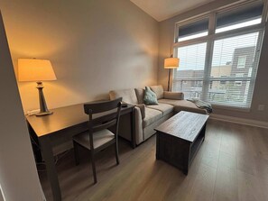 A modern living room with a sofa, a coffee table, a desk with a lamp, and a window with blinds. - Modern 1BR Bridge Park in Dublin with Balcony and Pool (Dublin)