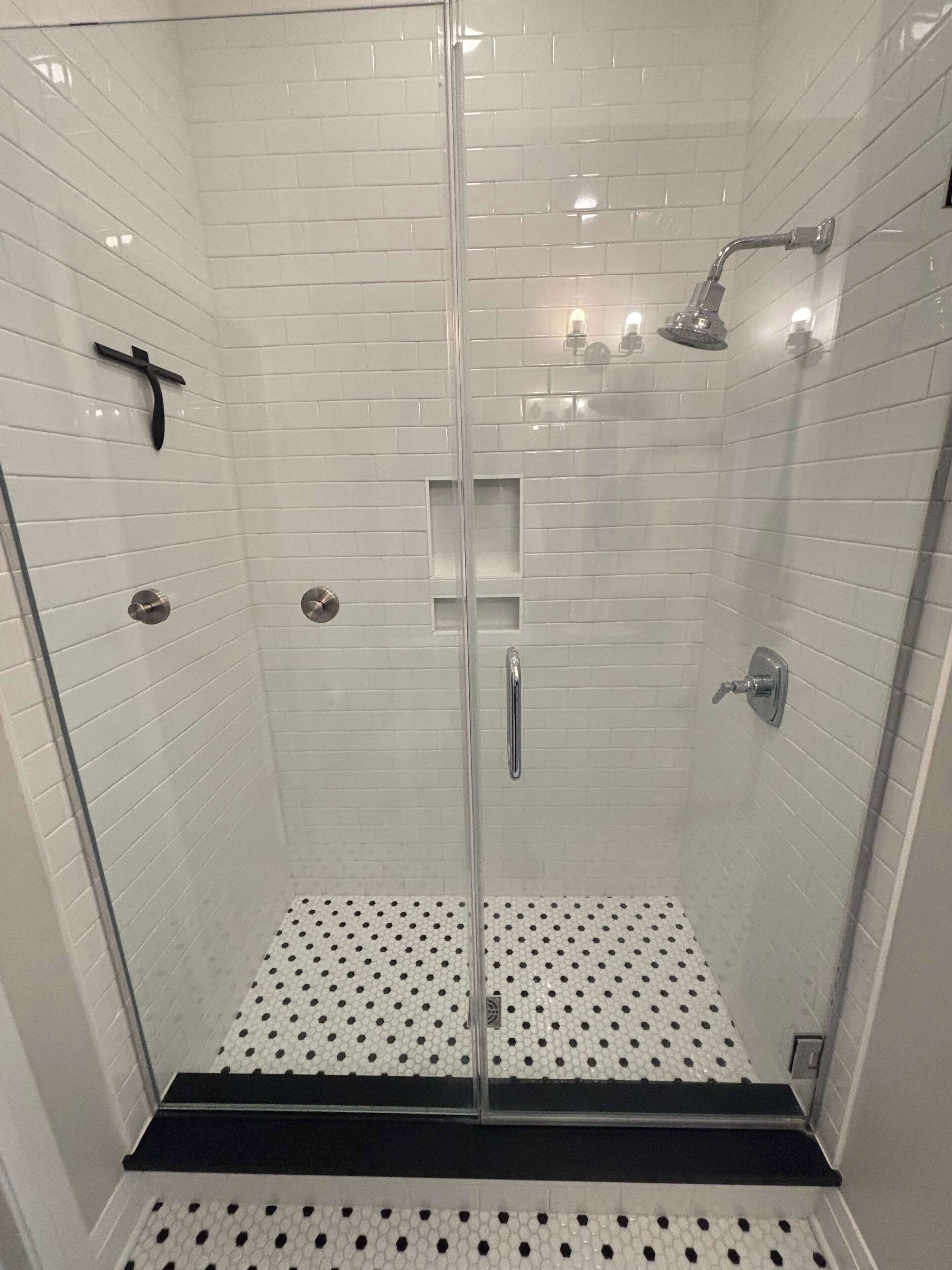 Une douche fermée par des parois de verre, avec un intérieur carrelé de carreaux blancs, une douchette à main et un robinet à main.