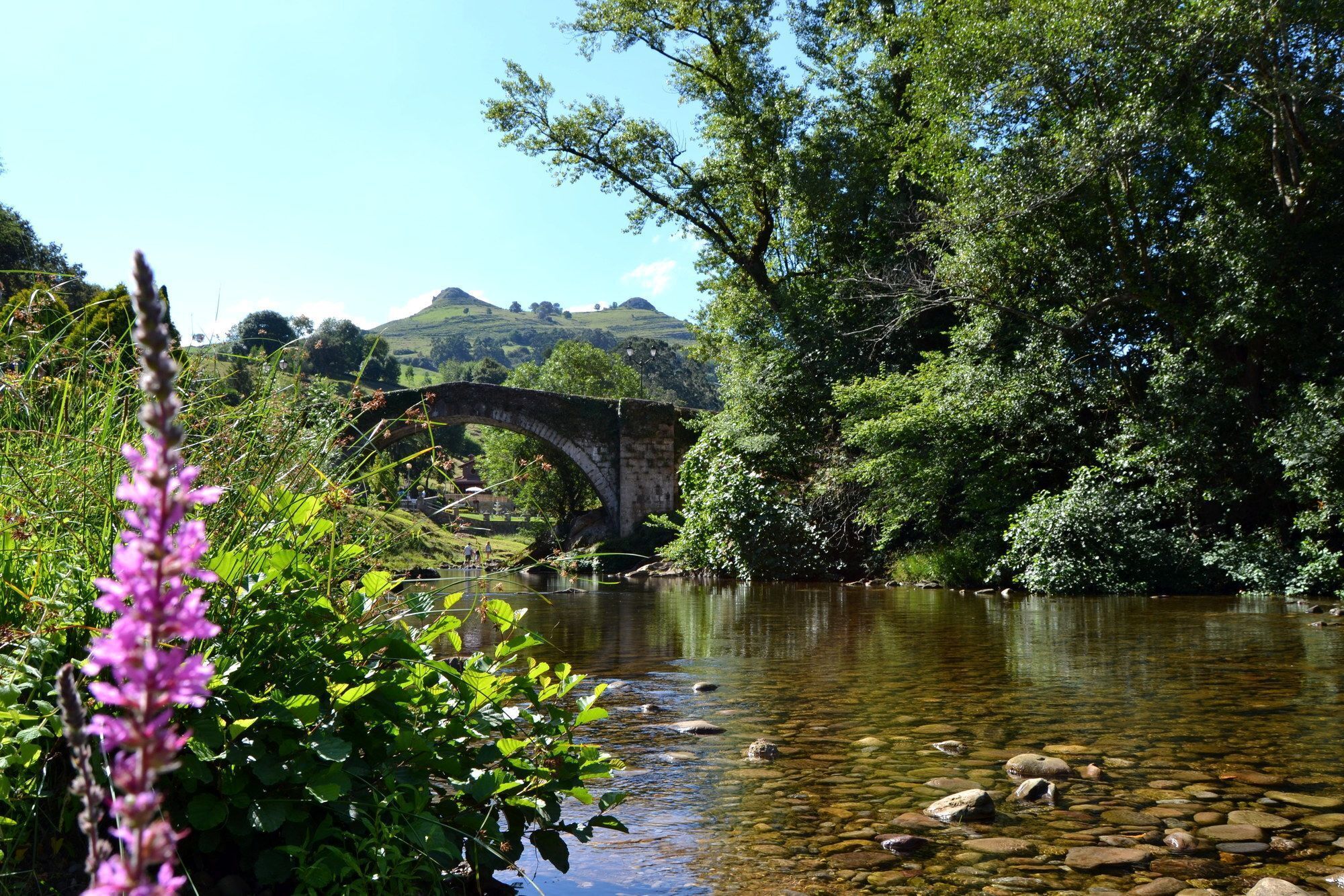 A stone bridge over a river with surrounding greenery and hills in the background.