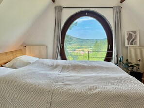 Ein Schlafzimmer mit großem Fenster, einem Bett mit strukturierter Decke, einem Heizkörper und einer Pflanze.