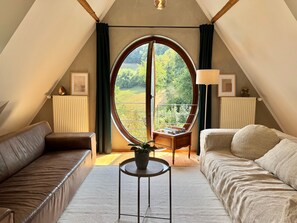 A cozy room with a large circular window, a sofa, a small table with a plant, and a lamp. - Apartment 'In den Bäumen' – Quiet Apartment on the Edge of the Forest near Freiburg (Bollschweil)