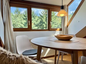A room with a round table, a wooden bowl, a white chair, and a view of trees through large windows. - Apartment 'In den Bäumen' – Quiet Apartment on the Edge of the Forest near Freiburg (Bollschweil)