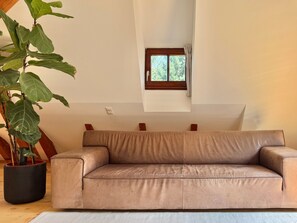 Un canapé beige dans une pièce agrémentée d’une grande plante en pot, d’une petite fenêtre et d’un plafond en bois.