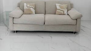 A light-colored sofa with two decorative pillows against a plain wall and a marble floor.