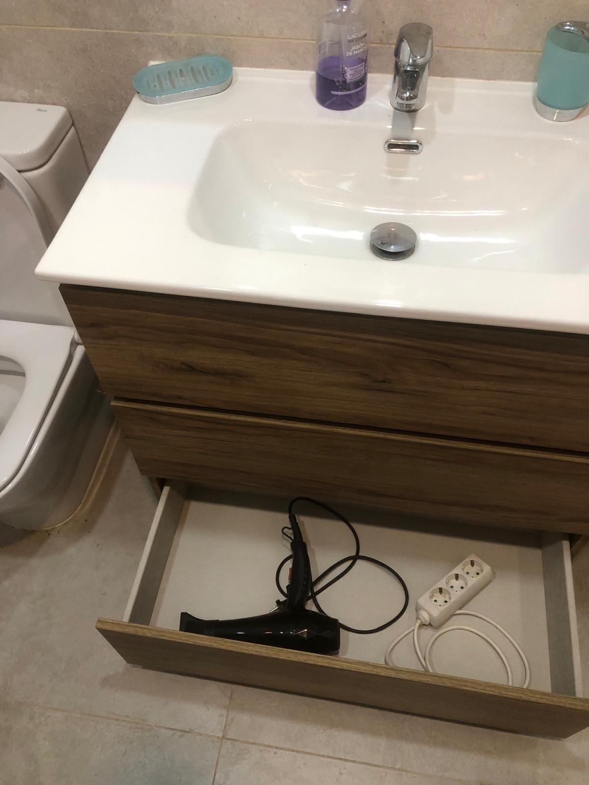 A bathroom with a white sink, wooden cabinet, and a drawer containing a black hairdryer and a white power strip.