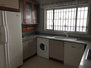 A compact kitchen with a refrigerator, washing machine, dishwasher, and a window with bars.