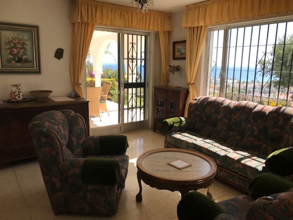 A living room with a sofa, armchair, and a round coffee table. There is a view through a window with curtains.