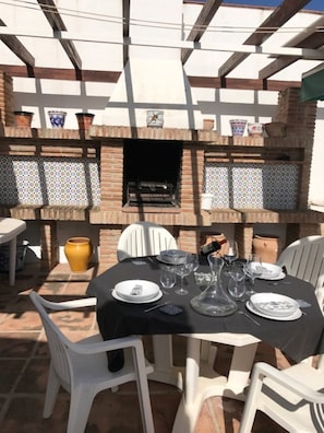 An outdoor dining area with a brick barbecue grill, white plastic chairs, and a table set for a meal.