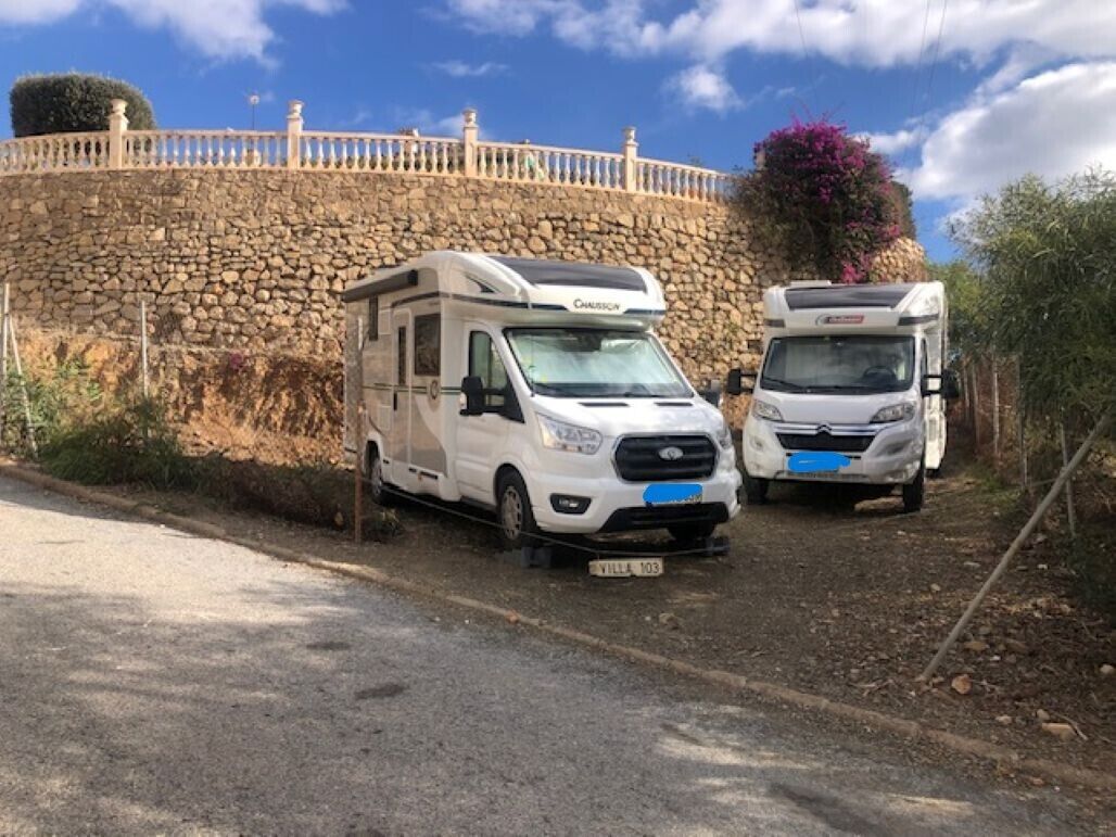 Two white motorhomes parked on a dirt road with a stone wall and a tree in the background.