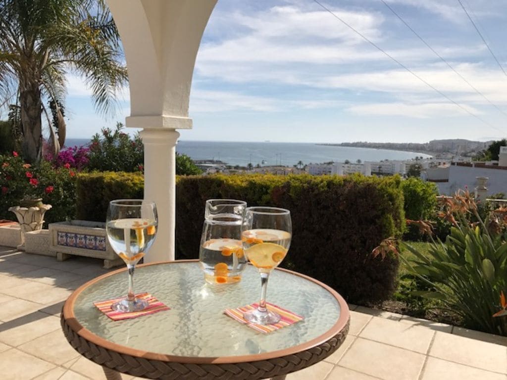 A glass table with two glasses of a clear beverage, a white column, and a view of the sea and buildings.