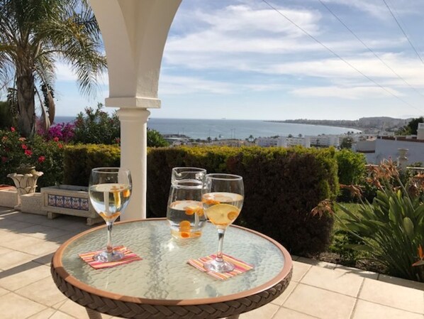 A glass table with two glasses of a clear beverage, a white column, and a view of the sea and buildings.