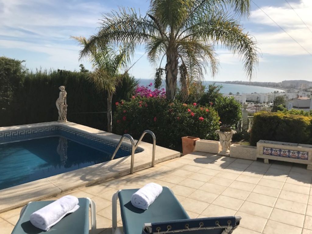 Un área de piscina con sillas de descanso y una estatua, rodeada de palmeras y flores.