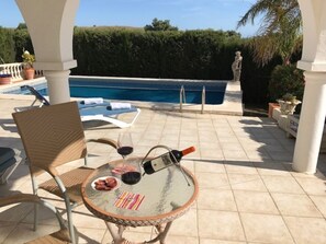 A glass-top table with a bottle and glasses, a chair, and a pool in the background.
