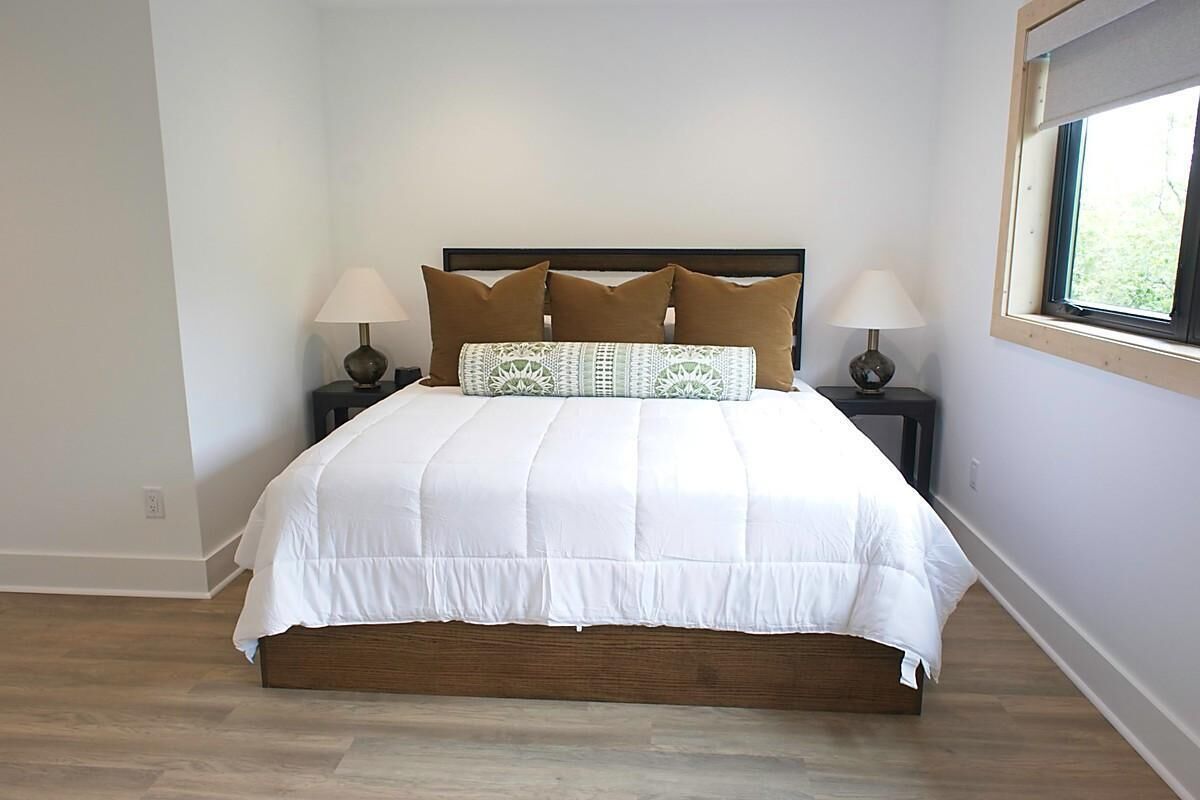 A neatly made bed with a white duvet and brown pillows, two bedside lamps, and a window with a view of greenery.