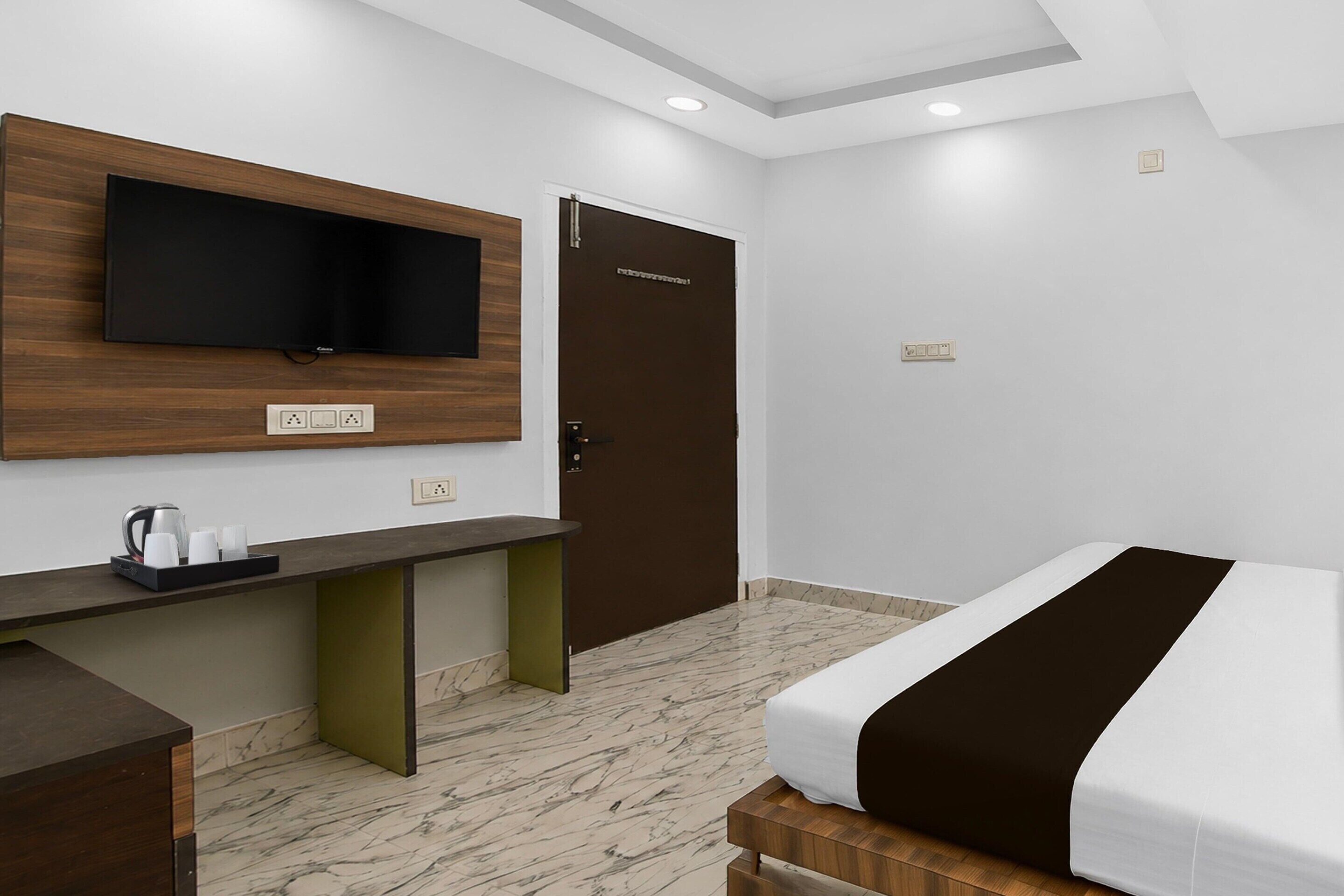 Una habitación de hotel con cama, escritorio con cafetera, televisor montado en la pared y puerta con picaporte.