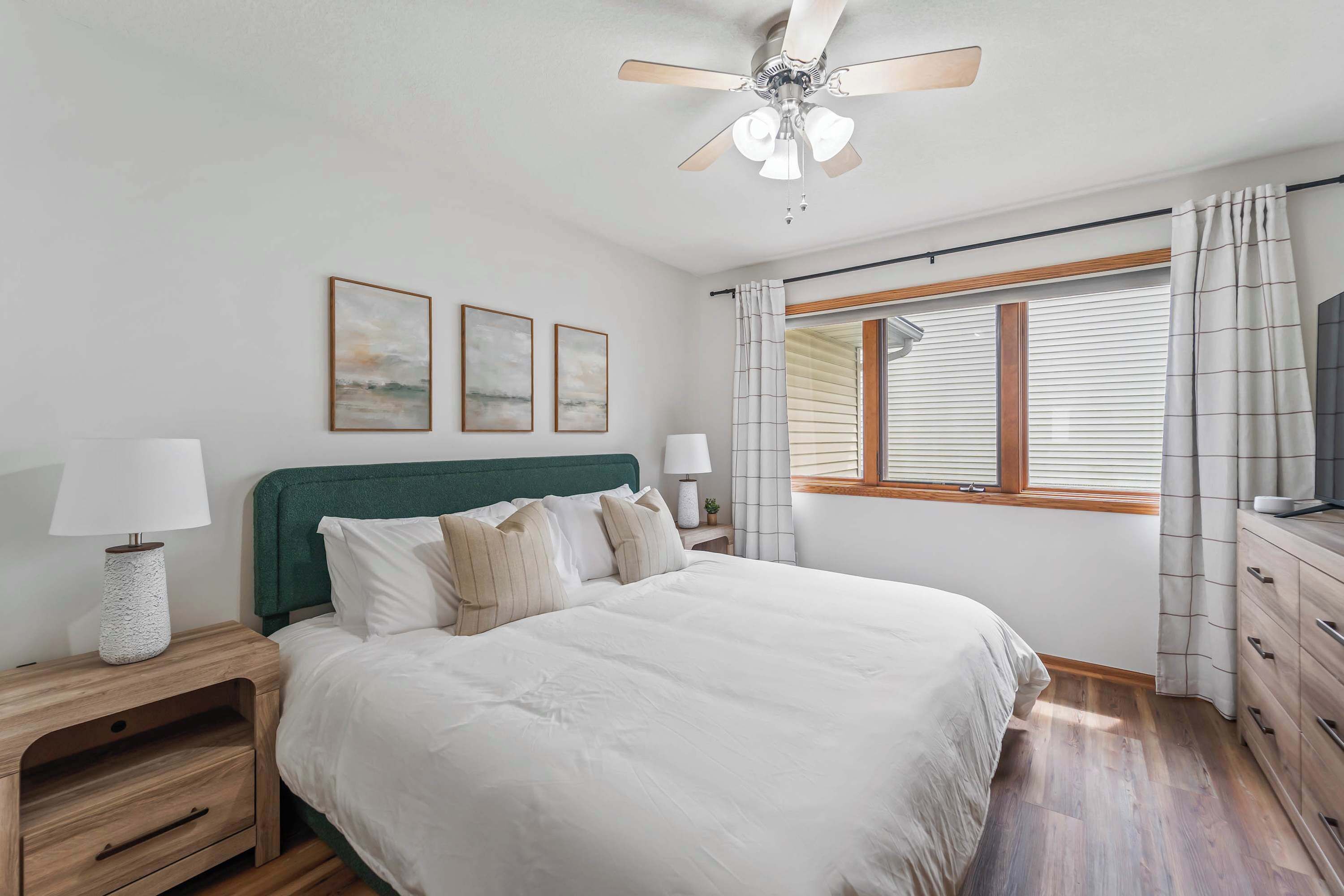 Un dormitorio con una cama, dos mesitas de noche, un ventilador de techo, una ventana con cortinas y tres cuadros enmarcados en la pared.
