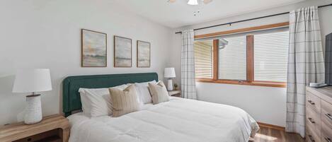 Un dormitorio con una cama, dos mesitas de noche, un ventilador de techo, una ventana con cortinas y tres cuadros enmarcados en la pared.