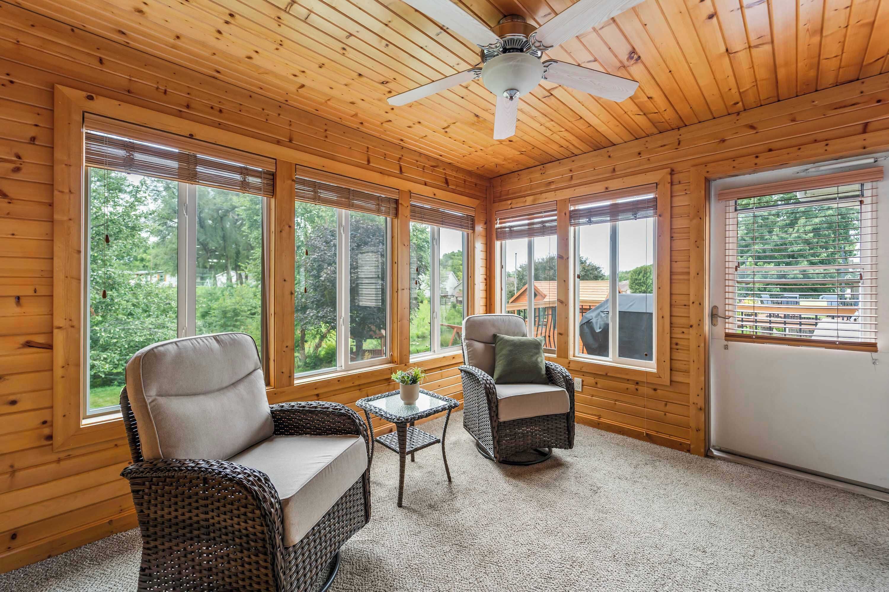 Une pièce aux murs et au plafond en bois, de grandes fenêtres, un ventilateur de plafond, deux fauteuils en osier, une petite table et une vue sur la verdure à l’extérieur.