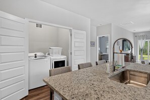 A modern kitchen with granite countertops, a stainless steel sink, and a dining area with a table and chairs. - Sea La Vie at OIB | Townhome Near Golf & Beaches (Ocean Isle Beach)