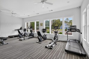 Ein Fitnessraum mit unterschiedlichen Trainingsgeräten, großen Fenstern und einem Deckenventilator.