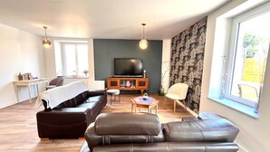 A modern living room with a sofa, ottoman, and a coffee table. There is a television on a wooden cabinet, and a window with a view. - Les Pierres de Lamballe – Countryside near beaches (Lamballe)