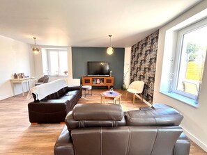 Ein modernes Wohnzimmer mit Sofa, Hocker und Couchtisch. Ein Fernseher steht auf einer Holzkommode, und ein Fenster bietet einen Ausblick.