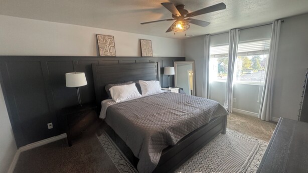 A bedroom with a bed, two bedside lamps, a ceiling fan, and a large window with curtains. - Family and pet friendly 5-bedroom home in charming Clearfield (Clearfield)