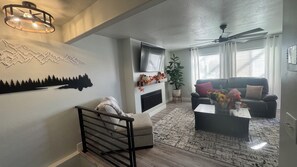 Ein Wohnzimmer mit Kamin, einem Wand-Fernseher, Deckenventilator und einem großen Fenster mit Jalousien.