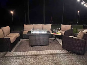 Ein Außenbereich mit Sitzgelegenheit, einem Tisch mit Glasplatte, gepolsterten Stühlen und einem Feuerelement.