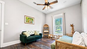 A room with a green sofa, a wooden chair, a ceiling fan, a window with curtains, and a giraffe decoration. - The Seaglass Bungalow - Where Colorful Comfort Meets Gulf Coast Charm (Gulfport)