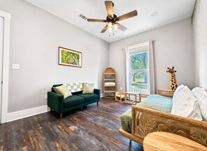 A room with a green sofa, a wooden chair, a ceiling fan, a window with curtains, and a giraffe decoration. - The Seaglass Bungalow - Where Colorful Comfort Meets Gulf Coast Charm (Gulfport)