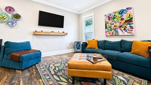 A living room with a flat-screen TV, a colorful abstract painting, a blue sofa, a brown ottoman, and a patterned rug. - The Seaglass Bungalow - Where Colorful Comfort Meets Gulf Coast Charm (Gulfport)
