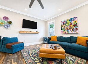 A living room with a flat-screen TV, a colorful abstract painting, a blue sofa, a brown ottoman, and a patterned rug. - The Seaglass Bungalow - Where Colorful Comfort Meets Gulf Coast Charm (Gulfport)