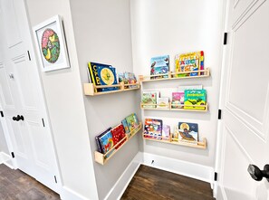 A hallway with white walls and wooden flooring. There are multiple wooden shelves mounted on the wall, each holding various children's books and educational materials. - The Seaglass Bungalow - Where Colorful Comfort Meets Gulf Coast Charm (Gulfport)