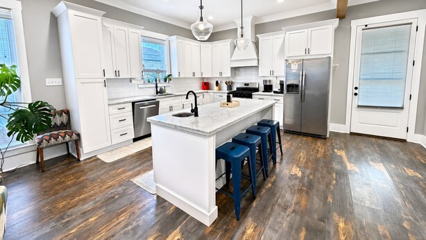 A modern kitchen with white cabinets, a central island, blue bar stools, and a stainless steel refrigerator. - The Seaglass Bungalow - Where Colorful Comfort Meets Gulf Coast Charm (Gulfport)