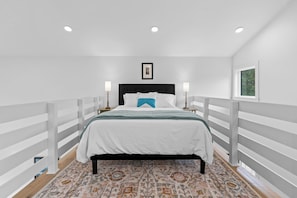 Un dormitorio con cama, dos mesitas de noche, un tapete estampado, un cuadro en la pared y una ventana.