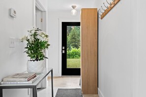 Un pasillo con pared blanca, una planta en maceta, una mesa con libros, una puerta que da a vistas de vegetación y un panel de madera.