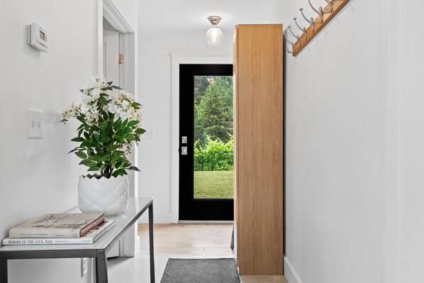 Un pasillo con pared blanca, una planta en maceta, una mesa con libros, una puerta que da a vistas de vegetación y un panel de madera.