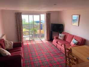 A living room with a red plaid carpet, a red sofa with patterned cushions, a television, and a dining table with chairs. - Tide Watch (Penzance)