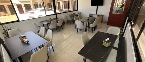 A modern dining area with multiple tables and chairs, large windows, and a flat-screen TV mounted on the wall.