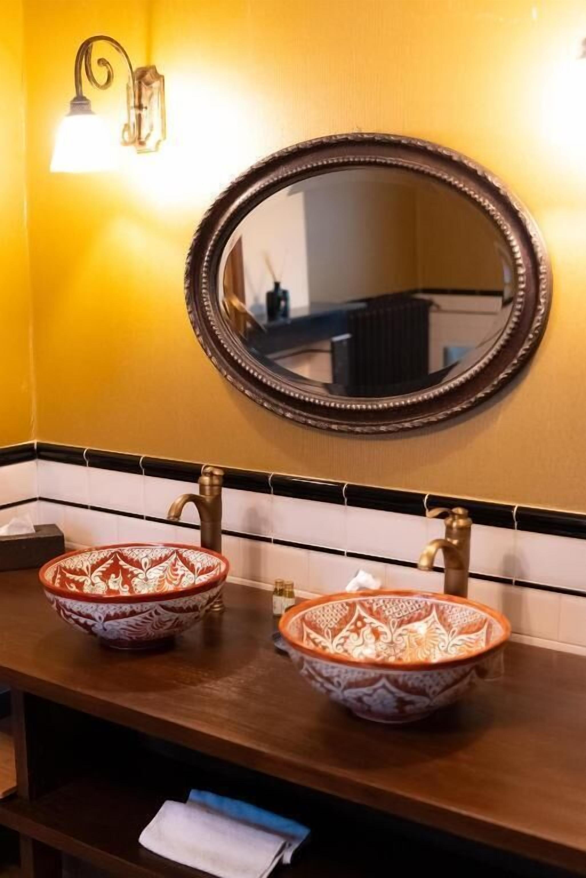 A bathroom with two ornate ceramic sinks, a wall-mounted oval mirror, and a light fixture above the mirror.