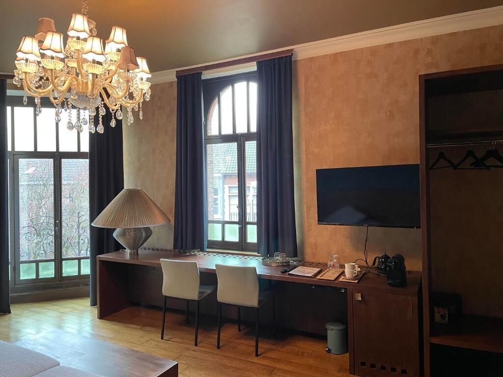 Una habitación con escritorio, sillas, televisor, candelabro y ventana con cortinas.